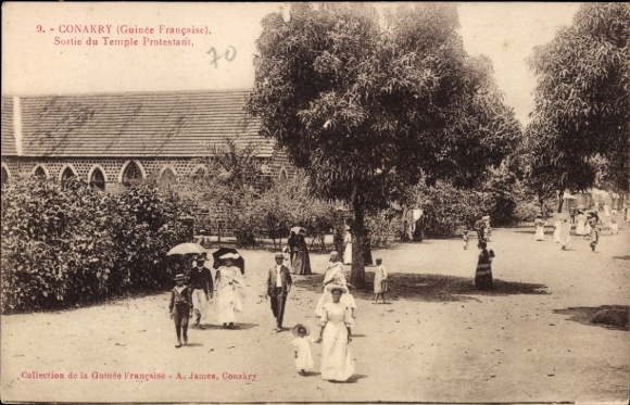 Ak Conakry Guinea, Menschen auf der Straße, Kirche im Hintergrund, Frauen in weißen Kleidern, Bäu